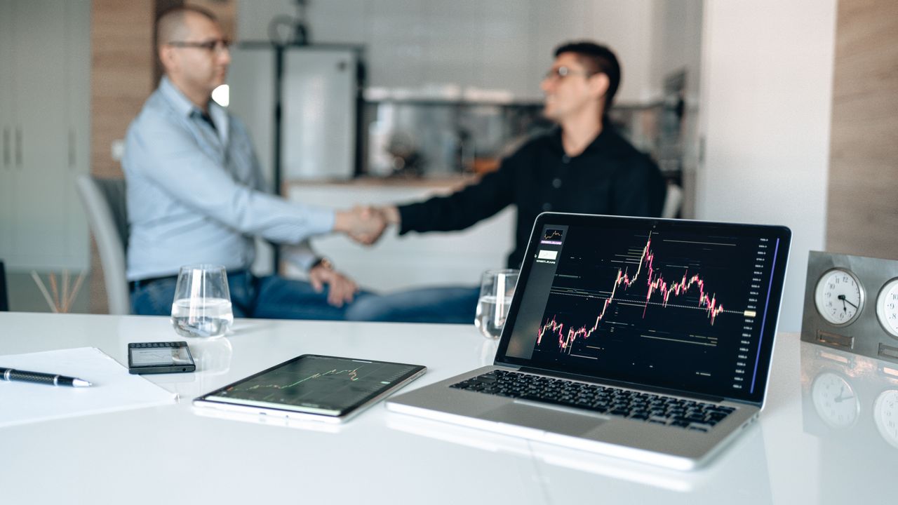 The Art of Drawing Readers In: Your attractive post title goes here Two businessmen shake hands at a desk with stock market graphs on a laptop.